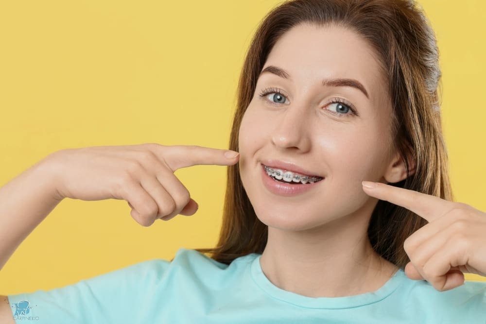 A smiling girl holds and points to an Invisalign Teen aligner with Carpinello Orthodontics in Drexel Hill, Newtown Square or Edgmont, PA, on a blue background.